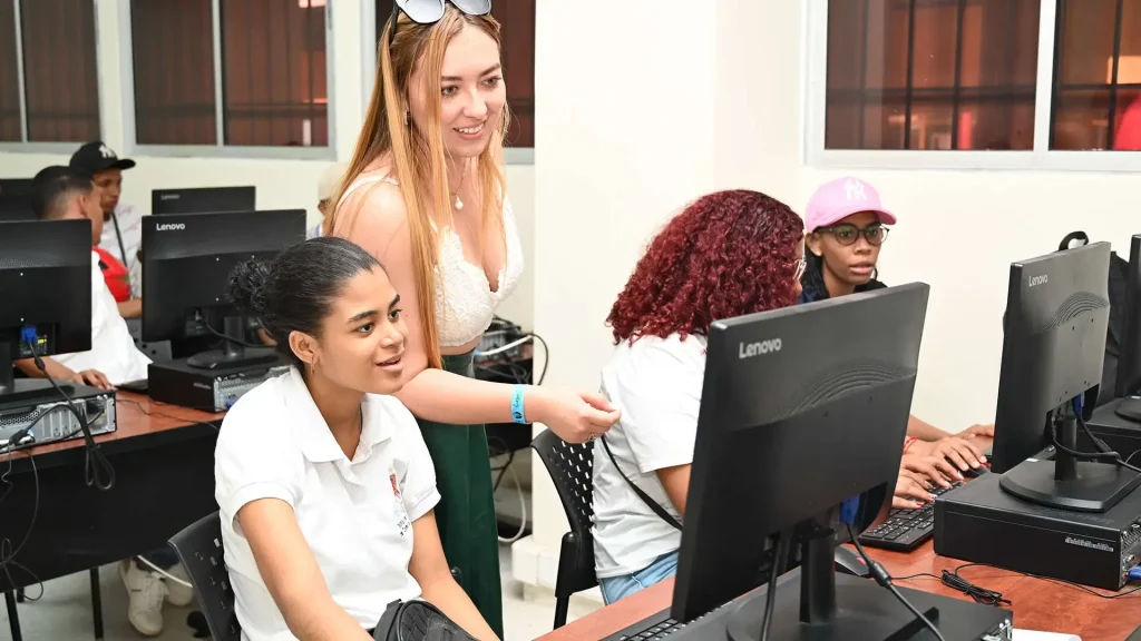 Una mujer rubia con blusa blanca ayuda a una estudiante frente a una computadora en un aula de informática, mientras otros alumnos trabajan en sus propios equipos.