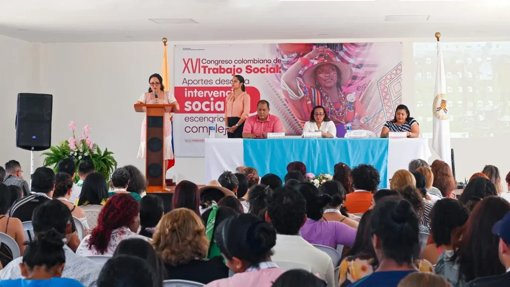 Un grupo de personas sentadas, escuchando a una mujer que habla desde un podio durante un evento. Se puede ver un cartel que dice "Congreso Colombiano de Trabajo Social".