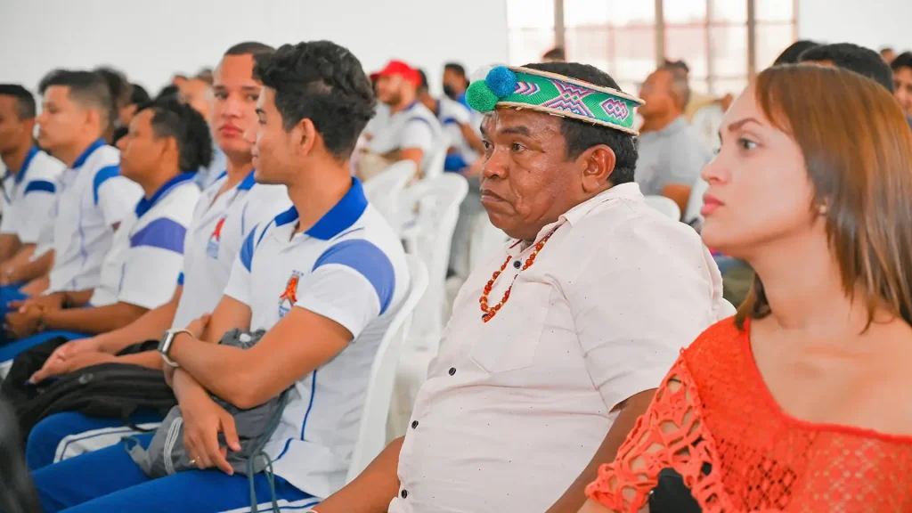 Hombres y mujeres en un auditorio. Resaltan hombres jóvenes con uniformes y un hombre con elemento en la cabeza, representativo de la cultura Wayuu.