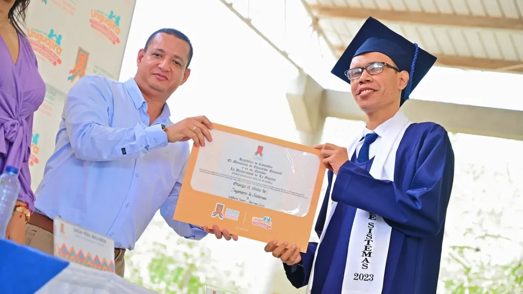 Un hombre vestido formal azul y otro con toga de graduación y lentes. Ambos están sosteniendo un diploma.