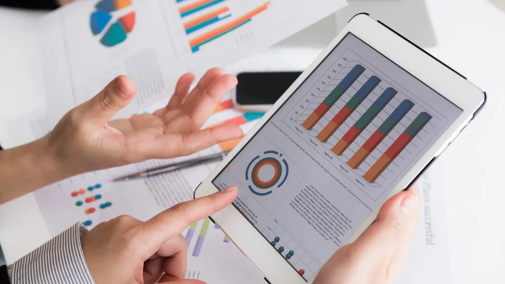 Primer plano de dos personas analizando gráficos y datos en una tablet y en documentos sobre una mesa.