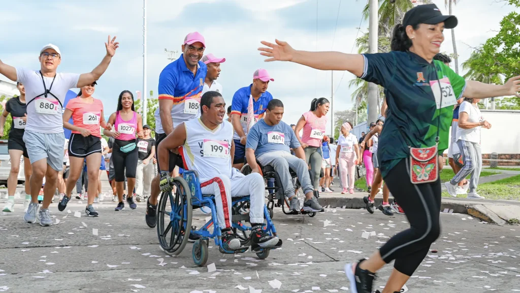 hombres y mujeres con los brazos arriba sonriendo y corriendo junto a un hombre empujando a una persona discapacitada en silla de ruedas en el desarrollo de la carrera.