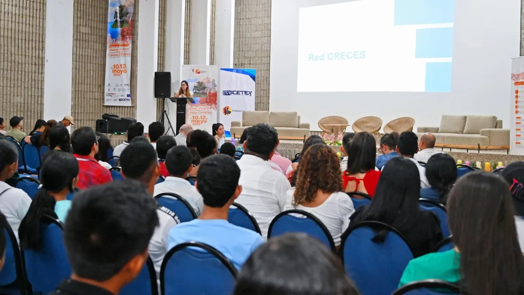 Mujer joven habla por un micrófono, frente al público grande de un auditorio.