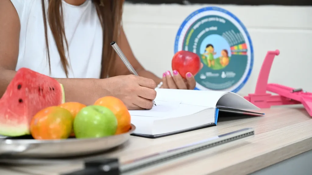 Una persona escribiendo en un cuaderno mientras sostiene una manzana, con fruta en la mesa.
