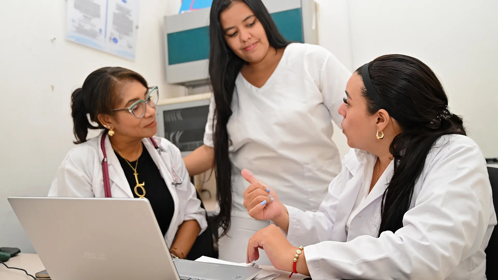 Tres mujeres con vestimenta alusiva a actividades de la salud. Están en un consultorio hablando entre ellas y al frente tienen un computador abierto. Tres mujeres con vestimenta alusiva a actividades de la salud. Están en un consultorio hablando entre ellas y al frente tienen un computador abierto.