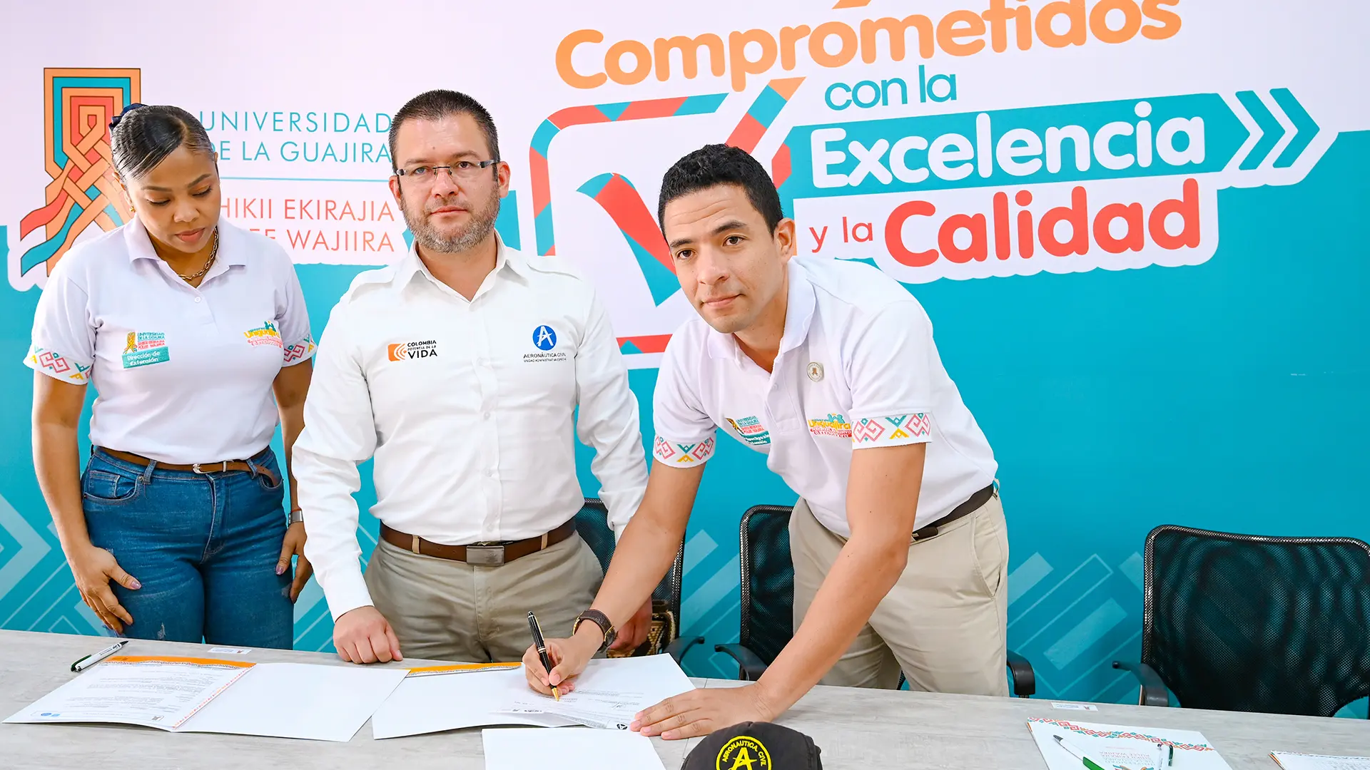 Dos hombres adultos firmando un documento al lado de un a mujer adulta