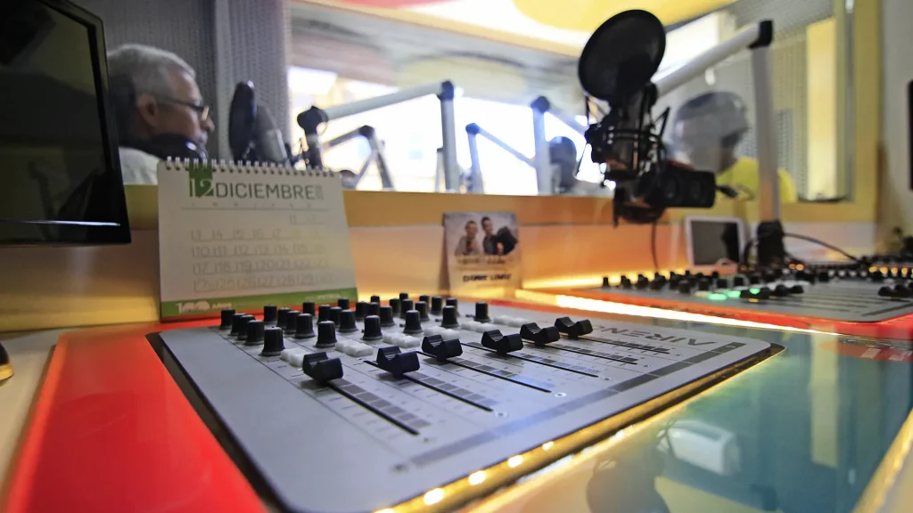 Primer plano de una mesa de mezclas en un estudio de radio, con un calendario en el fondo.