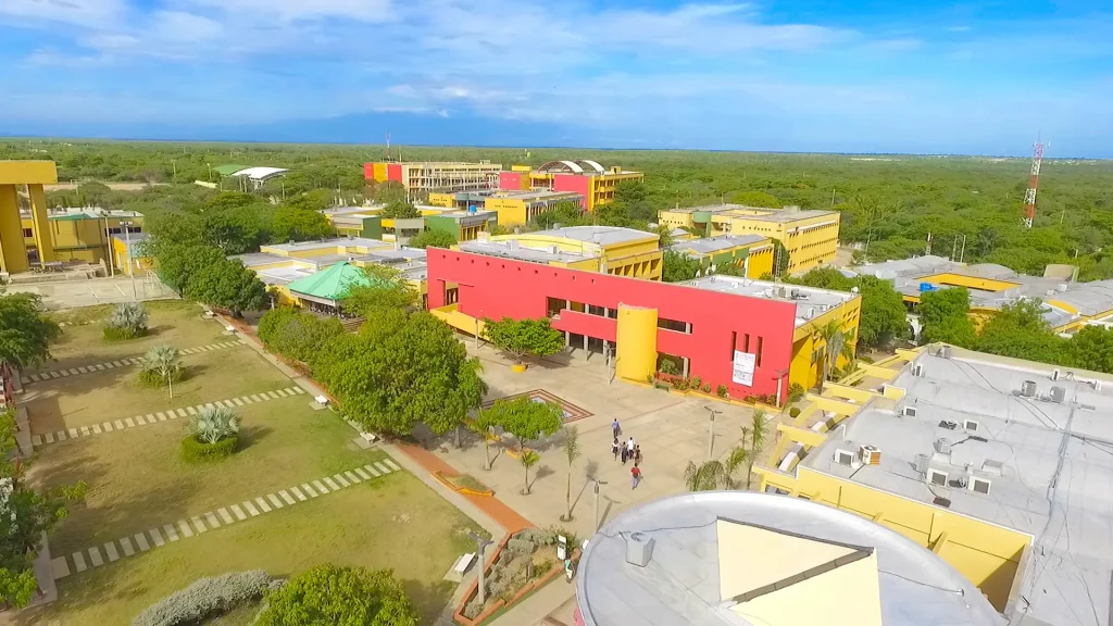 Vista aérea del campus de la Universidad de La Guajira, destacando un edificio central de color rojo y amarillo. Se aprecian amplias zonas verdes, senderos y personas caminando en el patio central.