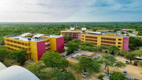Vista aérea del campus de la Universidad de La Guajira, mostrando varios edificios modernos de colores rojo y amarillo, rodeados de vegetación.