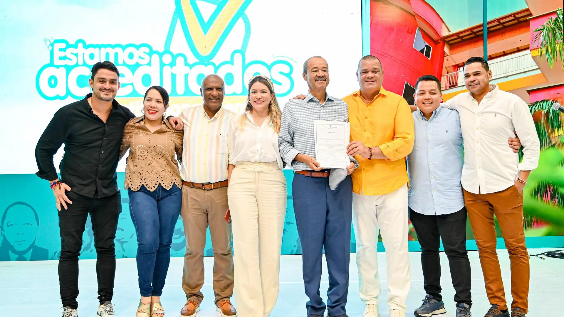 Un grupo de ocho personas posan juntas sonriendo en un escenario. En el centro, una persona sostiene un documento, mientras que los demás se agrupan a su alrededor de manera amigable. Detrás de ellos, hay una pantalla grande con un diseño que incluye la frase "Estamos acreditados" y elementos gráficos coloridos. El grupo está compuesto por hombres y mujeres de diferentes edades, vestidos con ropa casual y semiformal.