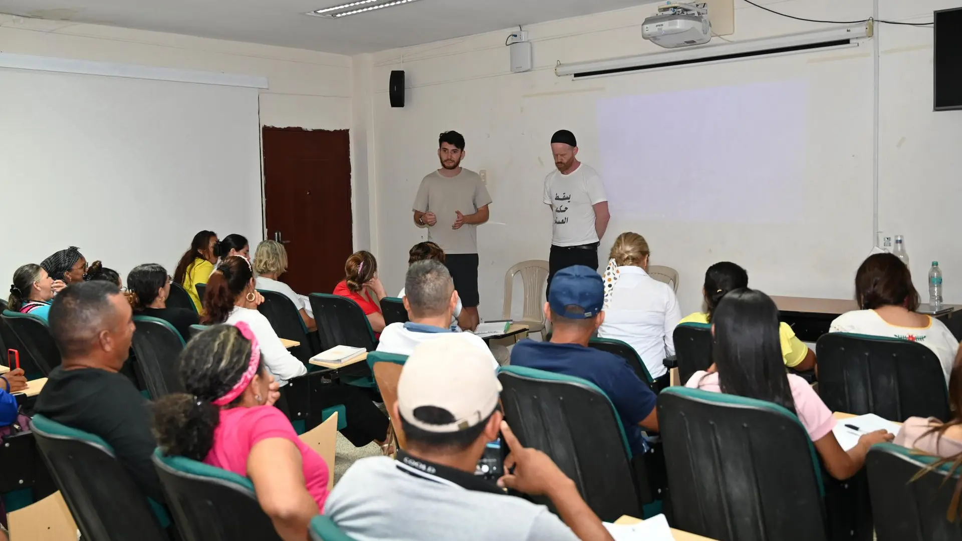 Un grupo de personas se encuentra en un aula de clases, sentadas y prestando atención a dos hombres jóvenes que están de pie al frente, dando una presentación. Los asistentes toman notas mientras observan la proyección en la pared. El ambiente es informal y participativo.