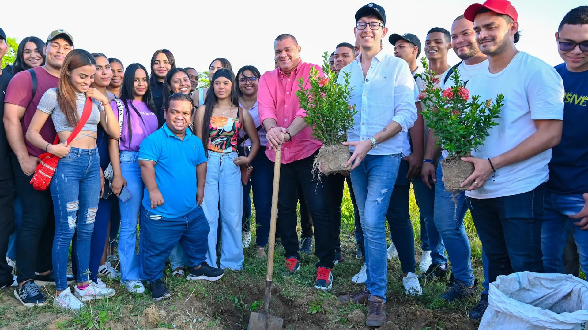Un grupo diverso de personas reunidas al aire libre en un entorno natural. Dos hombres al centro sostienen herramientas de jardinería y plantas listas para ser sembradas, mientras sonríen junto a otros participantes. La actividad parece estar relacionada con una iniciativa ambiental o comunitaria.