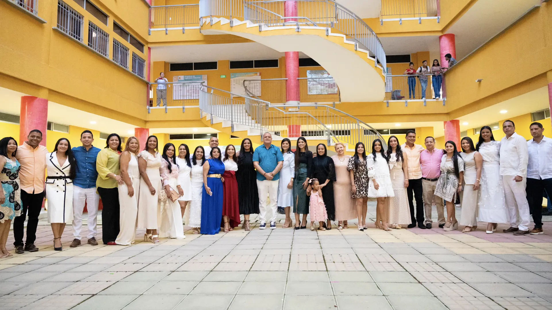 Grupo de personas posando sonrientes en el interior de un edificio de paredes amarillas y columnas rojas; todas visten ropa elegante y colorida, mientras al fondo se observa una escalera en espiral y algunas personas en el segundo piso.