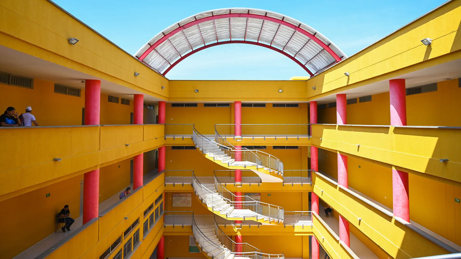 Vista interna de un edificio con paredes amarillas y columnas rojas. Las escaleras están dispuestas en un diseño moderno, con barandales metálicos curvos, y hay un techo semicircular translúcido que permite la entrada de luz natural. Varias personas están en los pasillos.