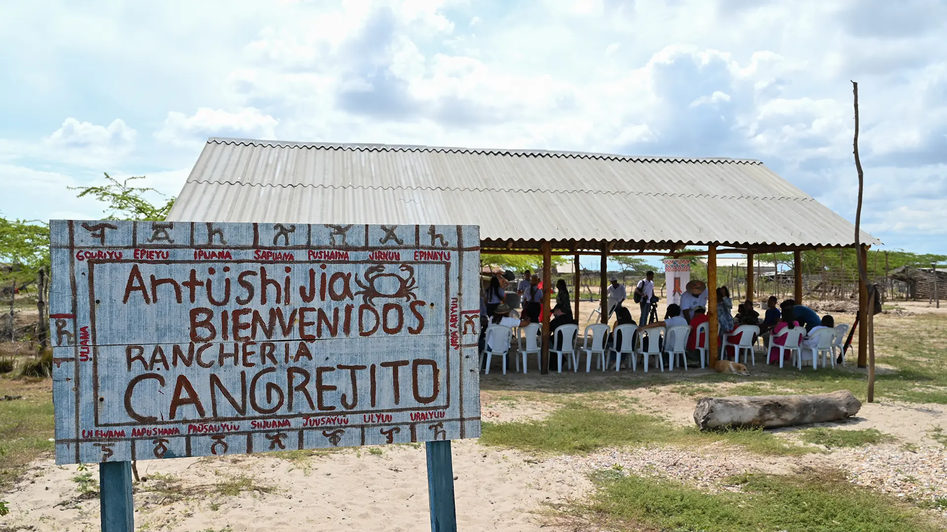 La composición de la imagen presenta un espacio abierto donde un grupo de personas se reúne bajo una estructura cubierta. El letrero en primer plano ocupa una parte considerable del espacio, y destaca por sus inscripciones en diferentes idiomas, presumiblemente, con la finalidad de indicar la bienvenida al lugar. La perspectiva es general, mostrando a las personas sentadas en sillas de plástico blancas, en el interior del cobertizo. En el fondo, se aprecian más estructuras y árboles dispersos, que dan contexto a la escena.