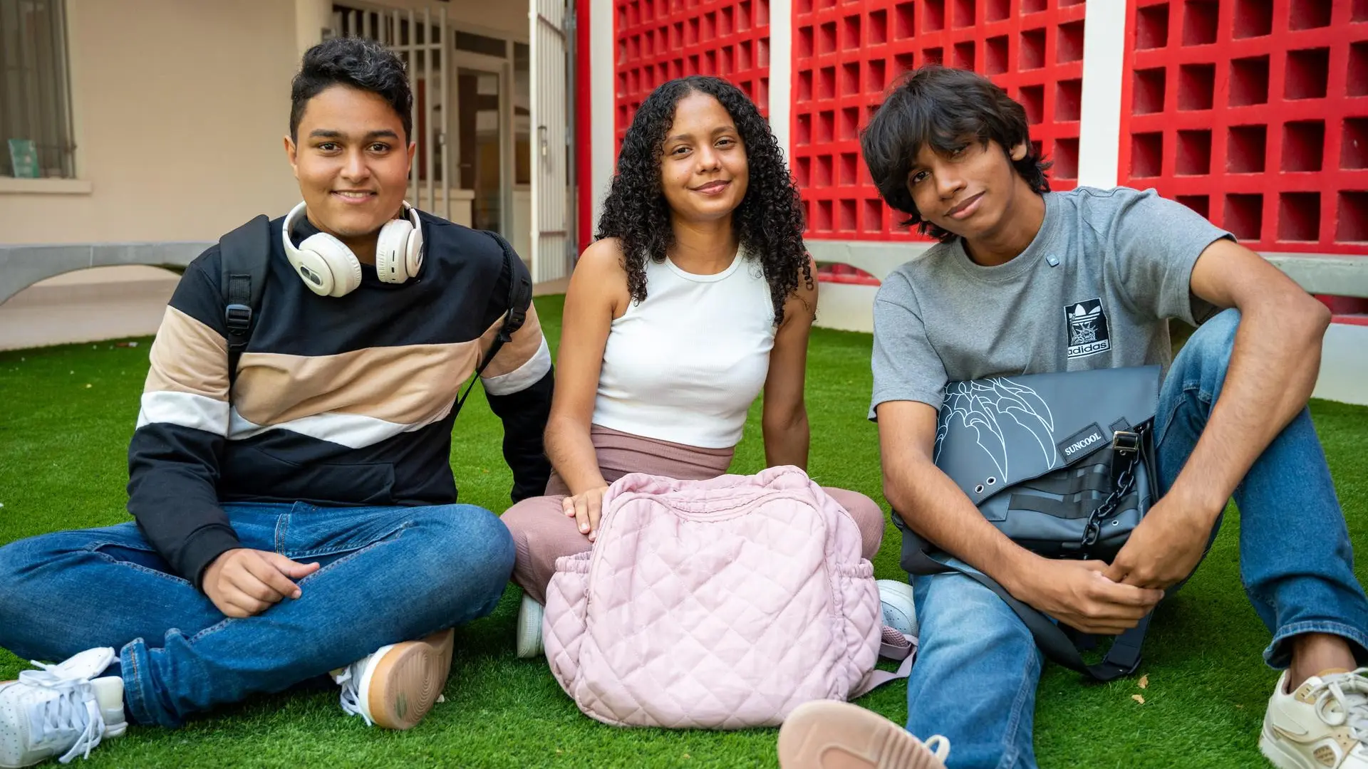 Tres estudiantes, dos hombres y una mujer, sentados sobre césped artificial en un campus universitario. Todos lucen relajados y sonrientes, con mochilas y audífonos, en un ambiente juvenil y acogedor con un fondo de paredes rojas y blancas.