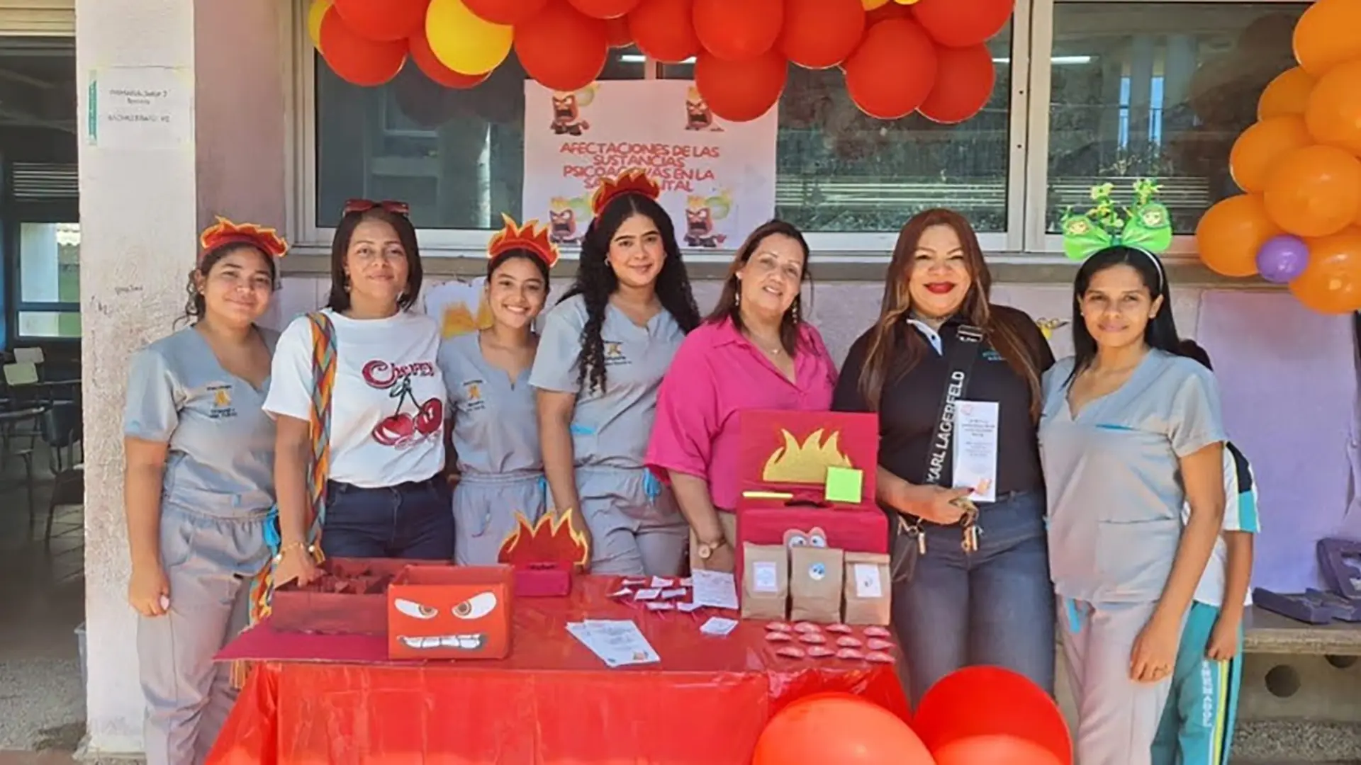 Grupo de mujeres, algunas con uniforme gris y otras con ropa casual, posan sonrientes frente a un stand decorado con globos rojos, naranjas y amarillos. El puesto está relacionado con la prevención de adicciones y tiene materiales informativos, adornos temáticos de fuego y carteles educativos sobre el impacto de las sustancias psicoactivas en la salud mental.