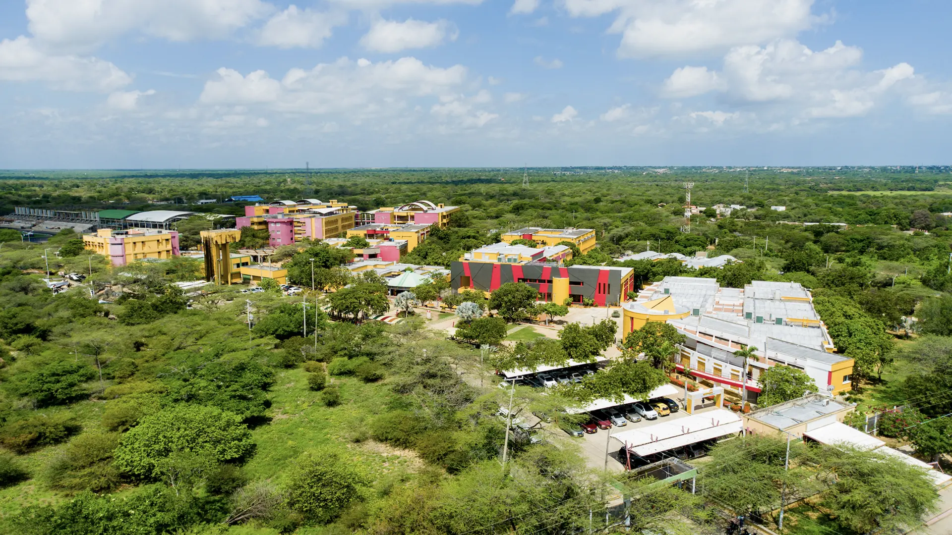 Vista aérea de un campus universitario rodeado de vegetación, con edificios de colores vivos como amarillo, rosa, rojo y negro, distribuidos entre zonas verdes y árboles, bajo un cielo parcialmente nublado.