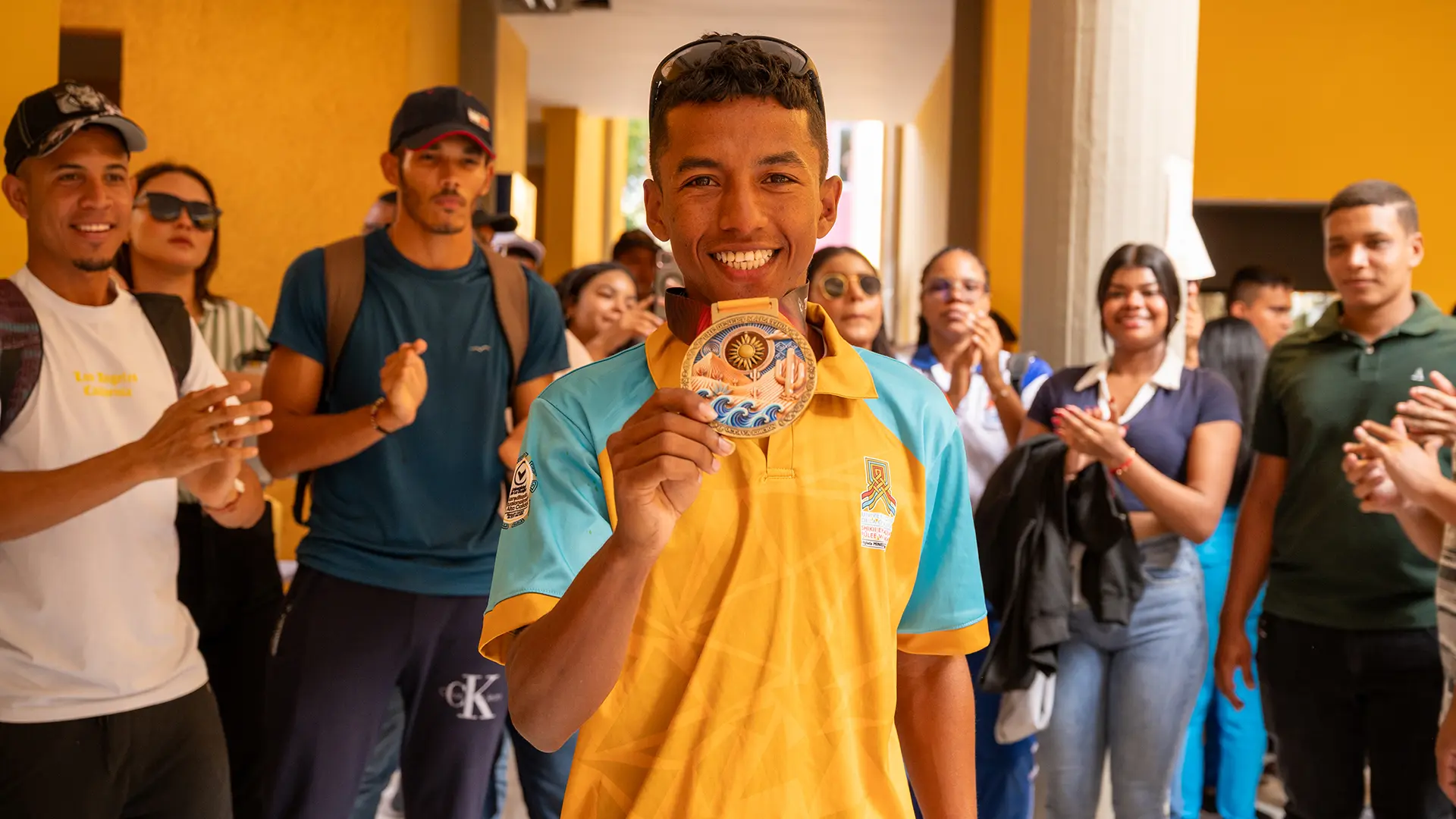 Joven deportista sonriente mostrando su medalla frente a un grupo de personas que lo aplauden. Viste camiseta deportiva amarilla y azul, y está rodeado de compañeros en un pasillo con paredes amarillas.