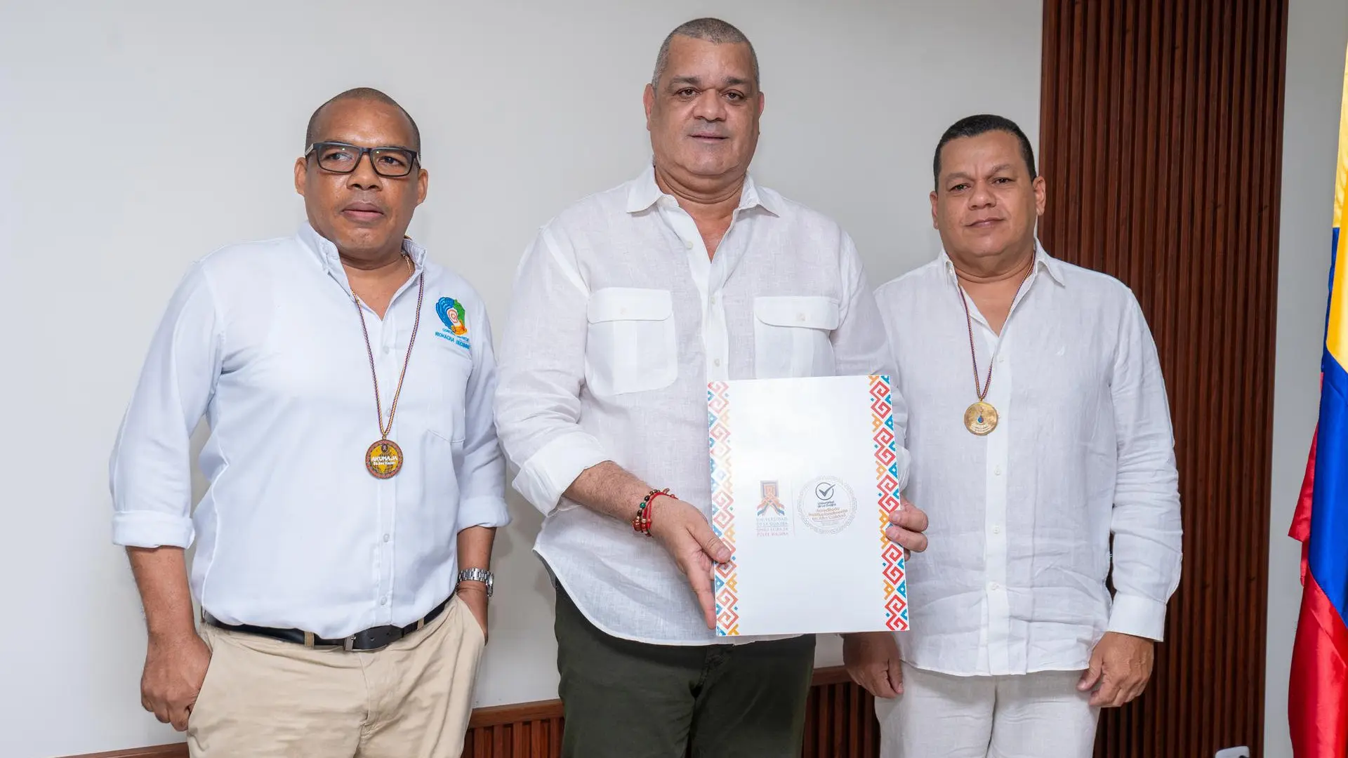Tres hombres posan juntos frente a una pared blanca. Todos visten camisas blancas de lino y llevan colgados del cuello medallones redondos. El hombre en el centro sostiene una carpeta blanca decorada con motivos étnicos en los bordes, la cual tiene logotipos institucionales. A la derecha se observa parcialmente una bandera de Colombia.