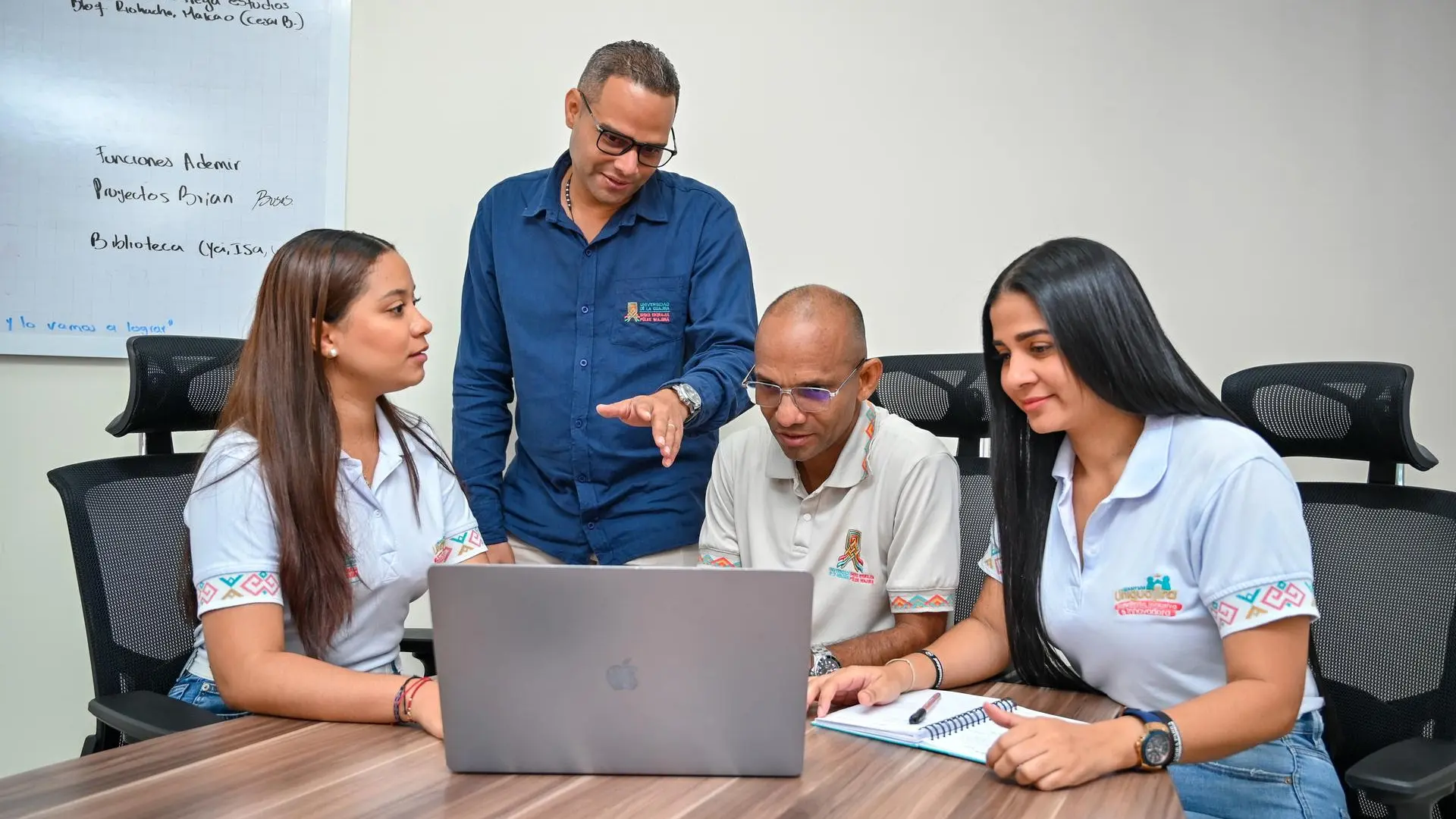 Cuatro personas, dos hombres y dos mujeres, se encuentran reunidas en una sala de reuniones observando una laptop. Parecen estar en una sesión de trabajo o capacitación, con una pizarra blanca al fondo y vestimenta institucional.