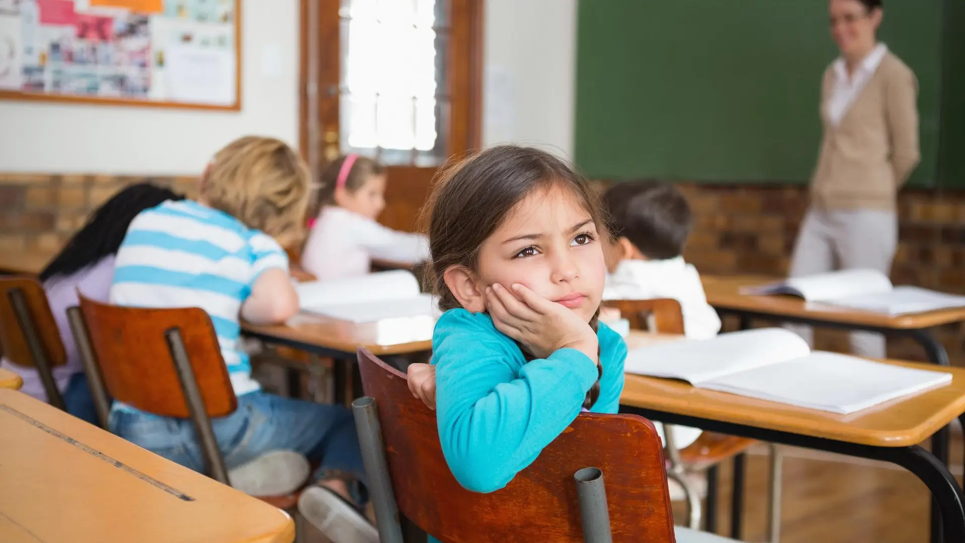 Una niña en edad escolar, con expresión pensativa y mirada perdida, se sienta en su pupitre con la cabeza recostada sobre la mano. A su alrededor, otros niños trabajan en sus cuadernos mientras una docente se encuentra de pie junto al tablero. La imagen transmite una sensación de distracción, desinterés o posible malestar emocional en un entorno de aula. Ideal para ilustrar temas relacionados con salud mental infantil o dificultades escolares.