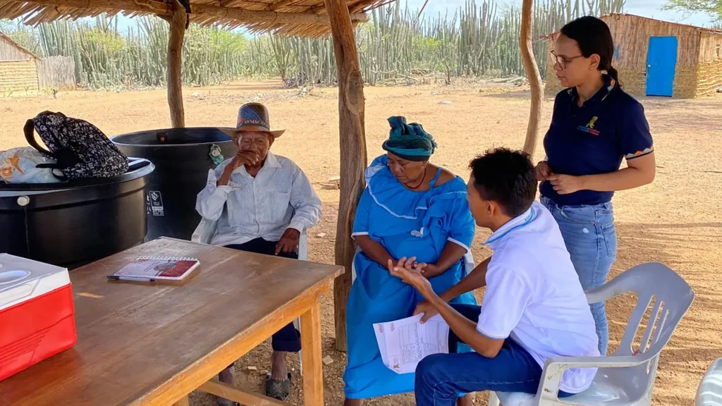 Dos adultos mayores indígenas conversan con dos personas jóvenes, bajo una choza de palma en una zona rural y árida, mientras revisan documentos. La escena refleja un proceso de consulta o atención comunitaria.