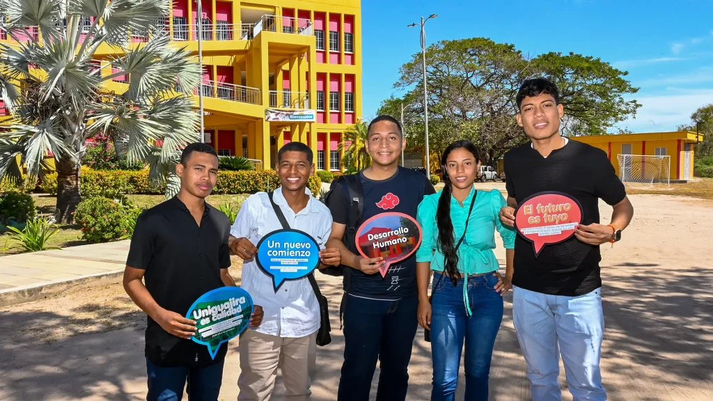 Grupo de tres estudiantes nuevos frente a la entrada principal de la sede Fonseca de la Universidad de La Guajira, sosteniendo carteles motivacionales.
