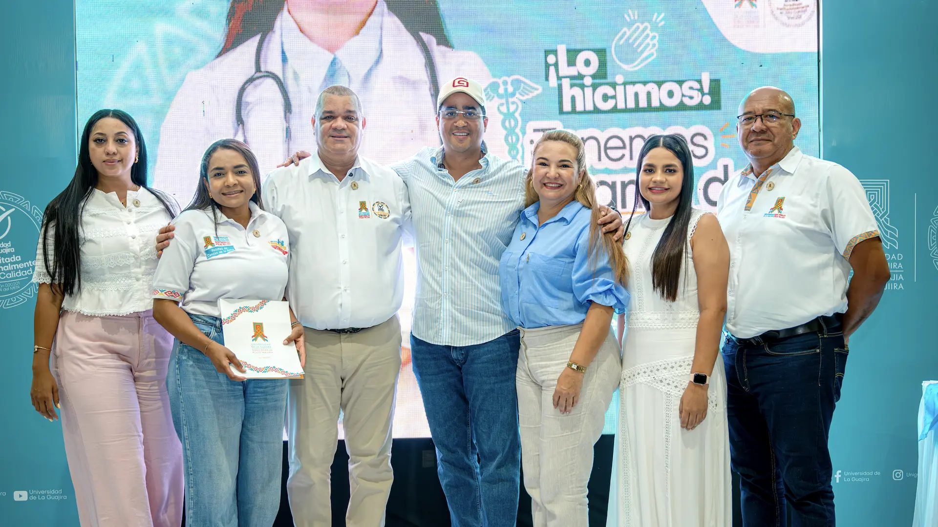 Grupo de siete personas posando frente a un fondo institucional de la Universidad de La Guajira; algunos visten camisas blancas con el logo de la universidad y otros ropa formal.