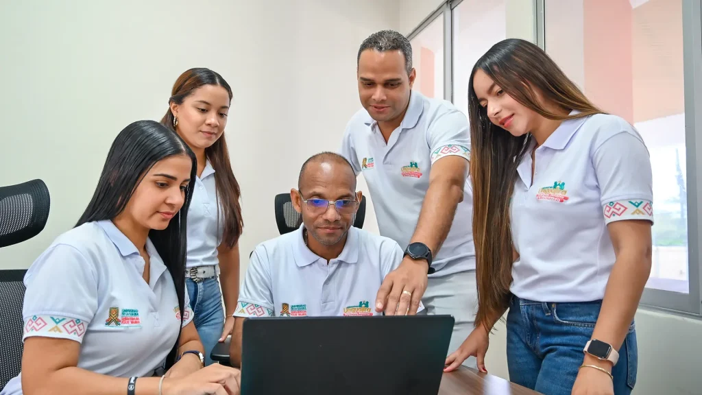 Cinco personas, tres mujeres y dos hombres, observan una pantalla de computador portátil mientras trabajan en equipo dentro de una oficina. Todos visten camisetas tipo polo blancas con el logo de Uniguajira.