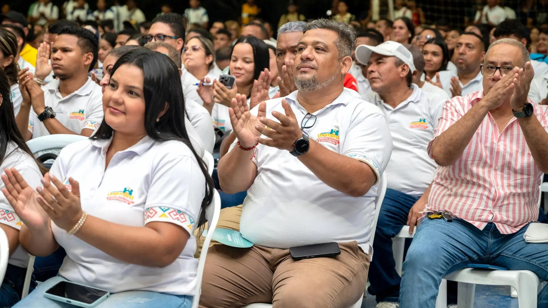 Público asistente a un evento de la Universidad de La Guajira, aplaudiendo desde sus asientos; la mayoría viste camisetas blancas con detalles coloridos en las mangas.