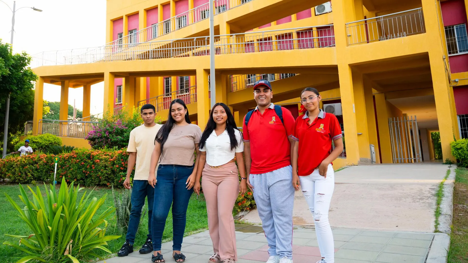 Cinco personas, entre estudiantes y personal, posan sonrientes frente a un edificio universitario amarillo con detalles rojos, en un entorno con jardines verdes.