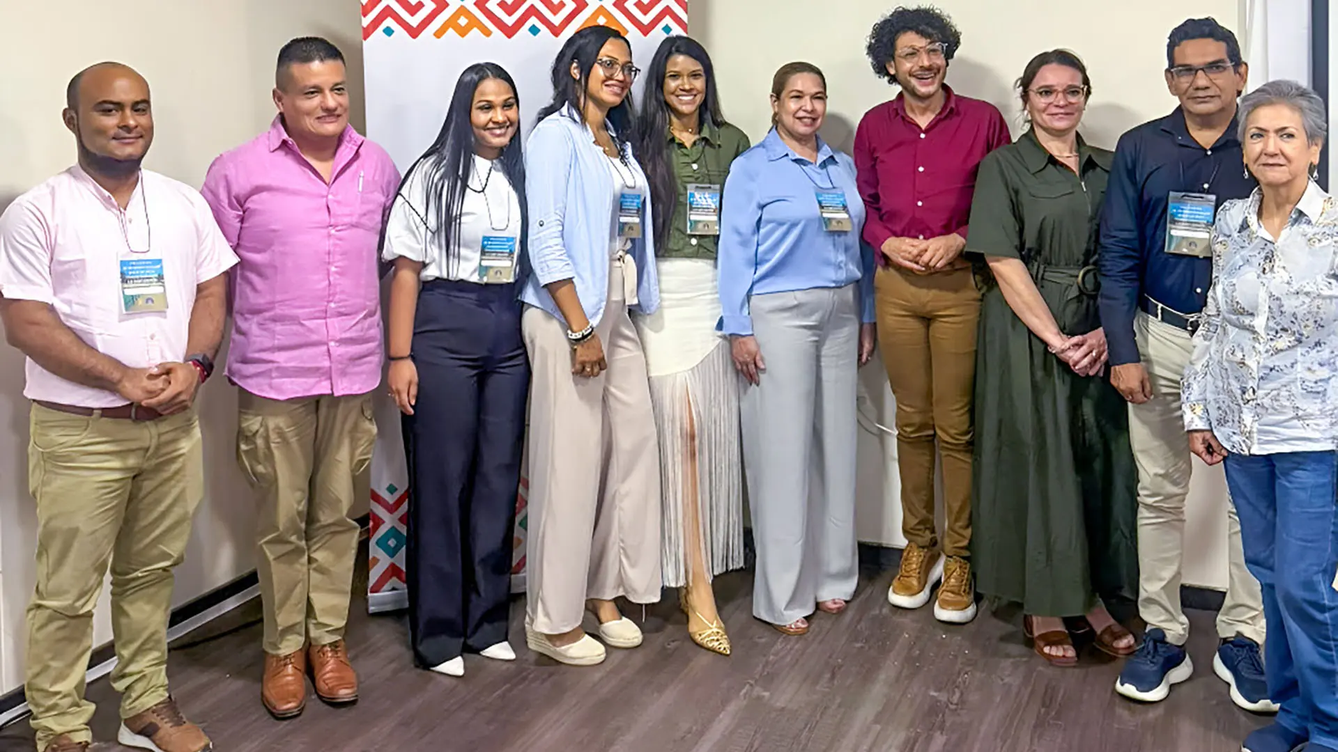 n grupo de hombres y mujeres posan juntos frente a un cartel con diseños coloridos en un espacio institucional. Todos llevan gafetes de identificación, lo que indica que participan en un evento académico o profesional.