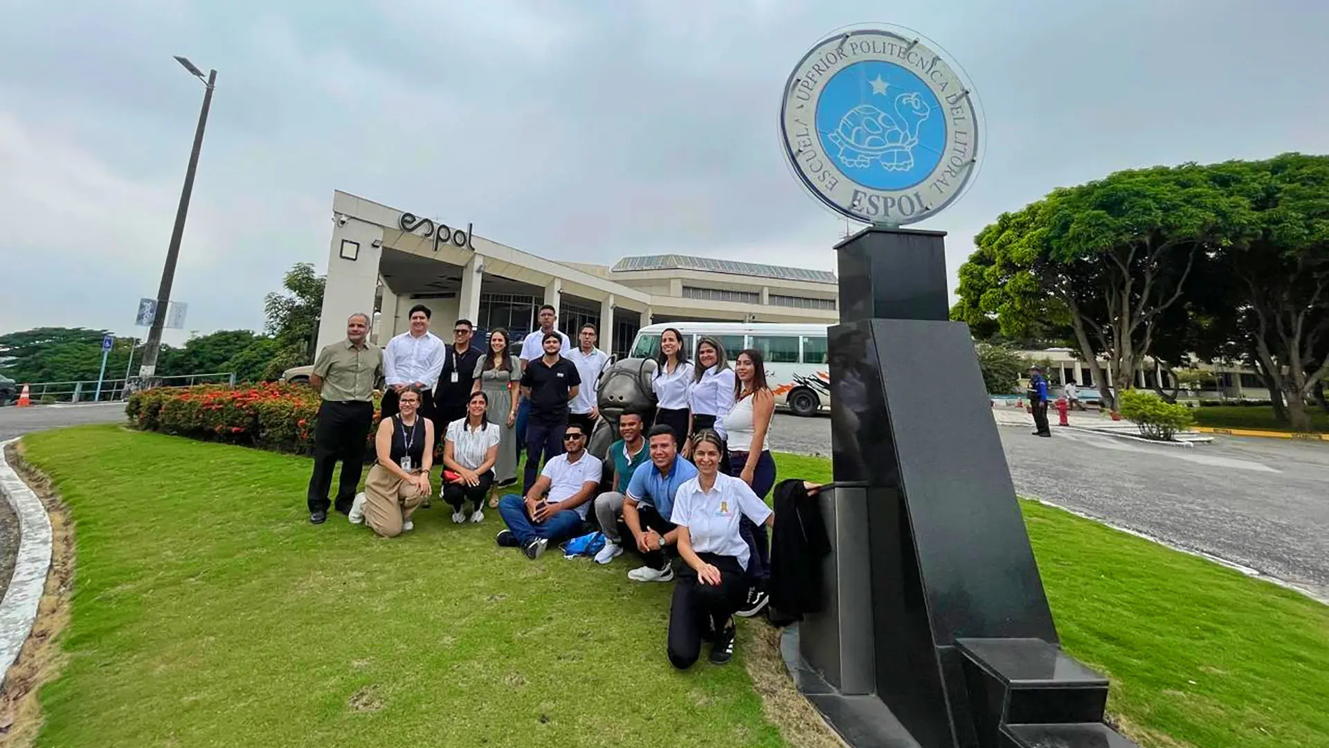 Grupo de estudiantes y docentes posan sonrientes frente a la entrada principal de la Escuela Superior Politécnica del Litoral (ESPOL), junto al monumento con el logo institucional.