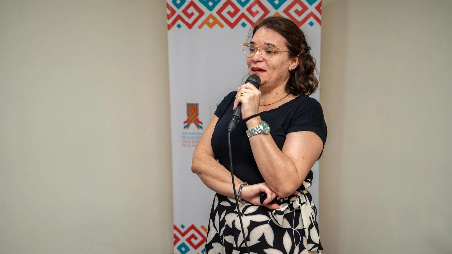 Mujer con gafas y blusa negra sostiene un micrófono mientras habla frente a un público, con un fondo de banner institucional de colores geométricos.