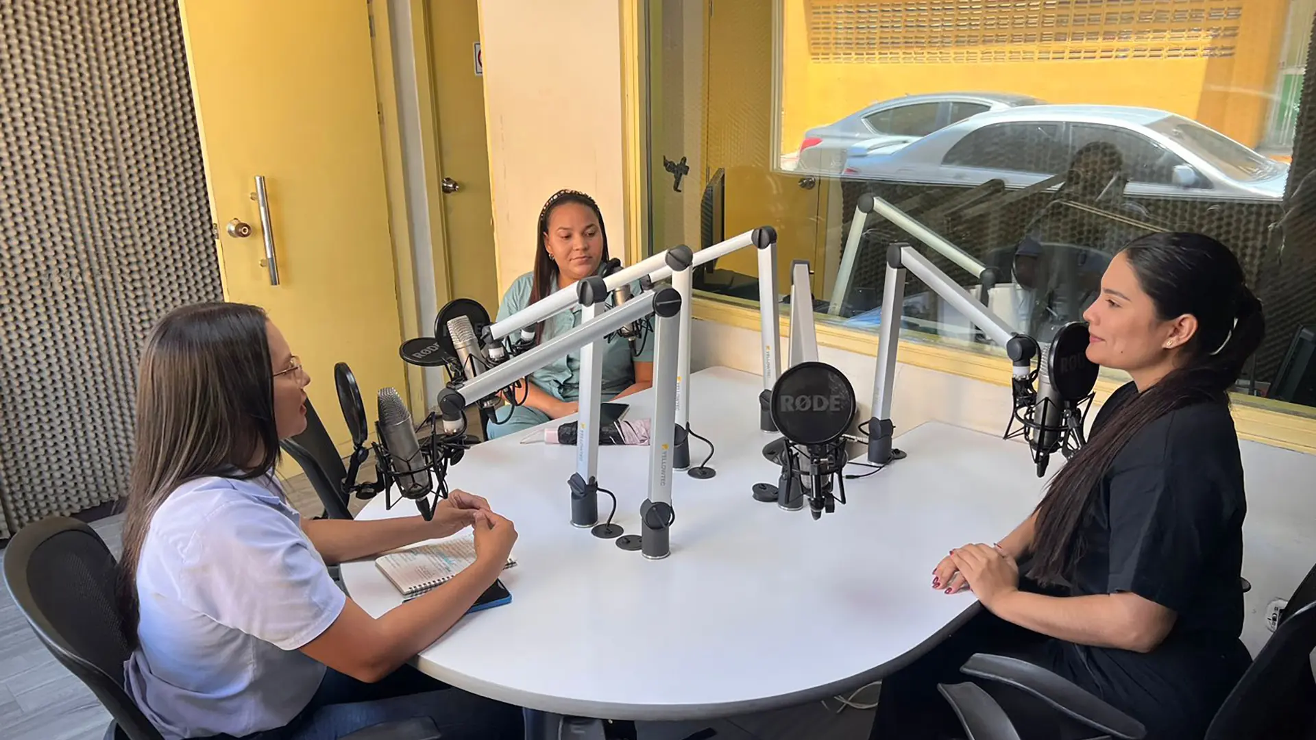 Tres mujeres participan en una entrevista radial en una cabina de radio. Están sentadas alrededor de una mesa circular con micrófonos profesionales. Una de ellas toma notas en un cuaderno mientras conversa con las otras dos.