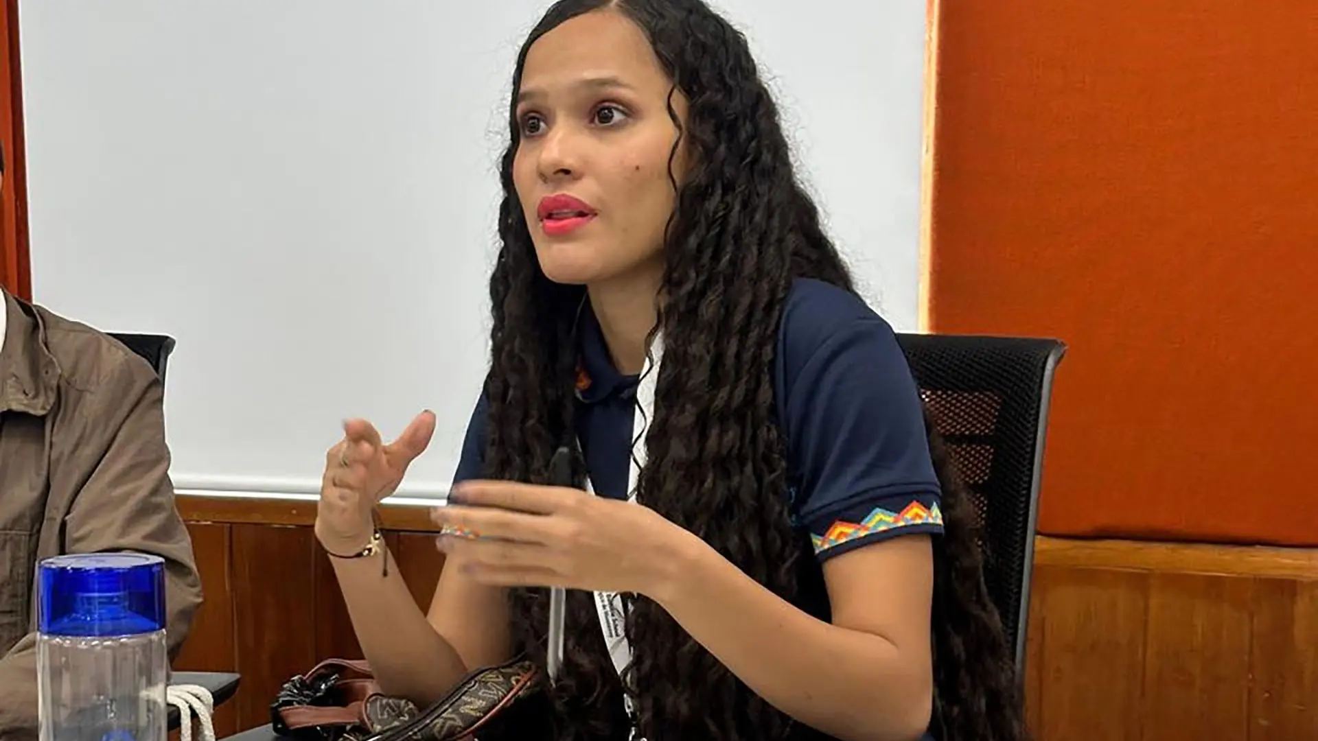 Joven estudiante participa activamente en una mesa de trabajo, gesticulando mientras interviene; frente a ella hay un cuaderno, un celular y un rótulo con su nombre.