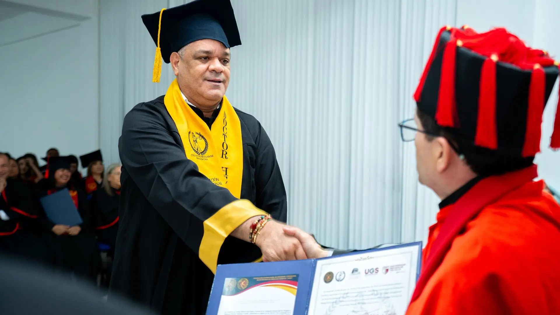 Hombre con birrete y banda amarilla que dice “Doctor HC” estrecha la mano de otro participante vestido con toga roja y negra durante una ceremonia de grado o reconocimiento académico.