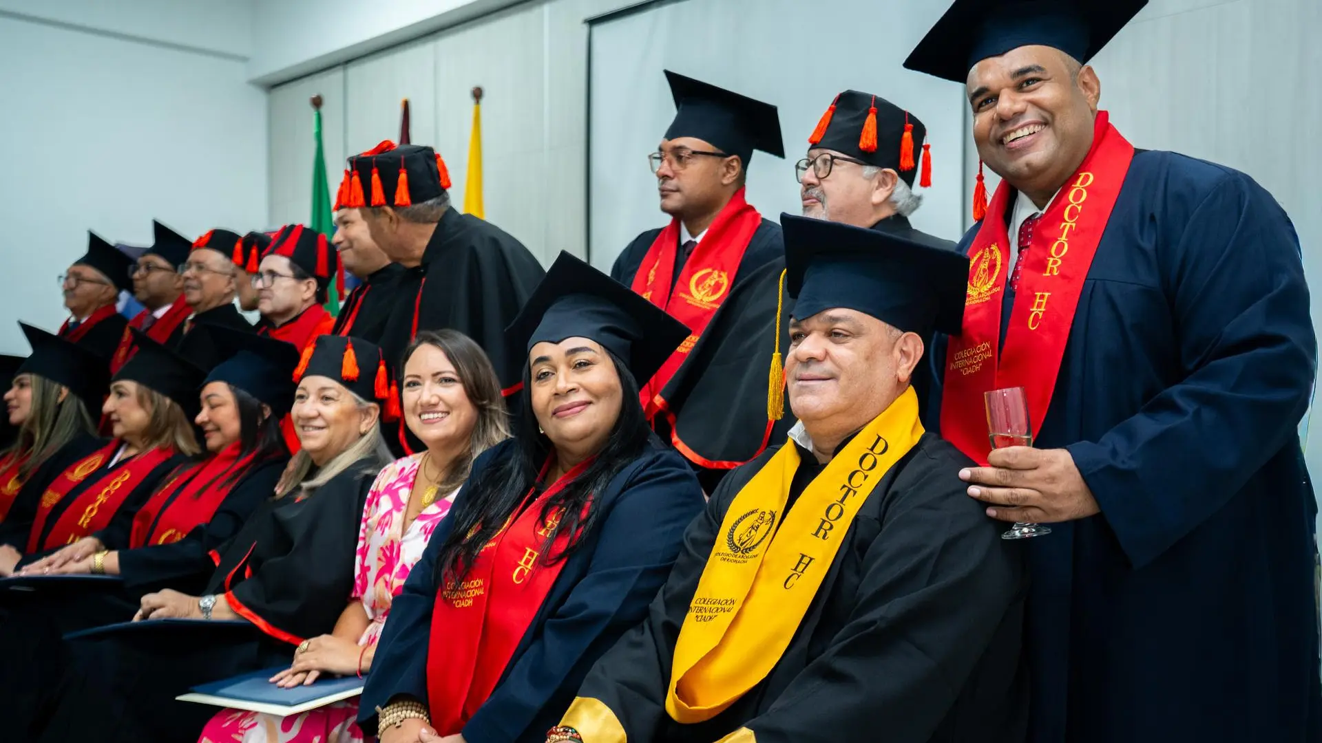 Grupo de personas vestidas con togas y birretes negros con detalles rojos posan para una fotografía grupal durante una ceremonia académica. En primera fila hay personas sonriendo, algunas con bandas rojas o amarillas que indican “Doctor HC”.