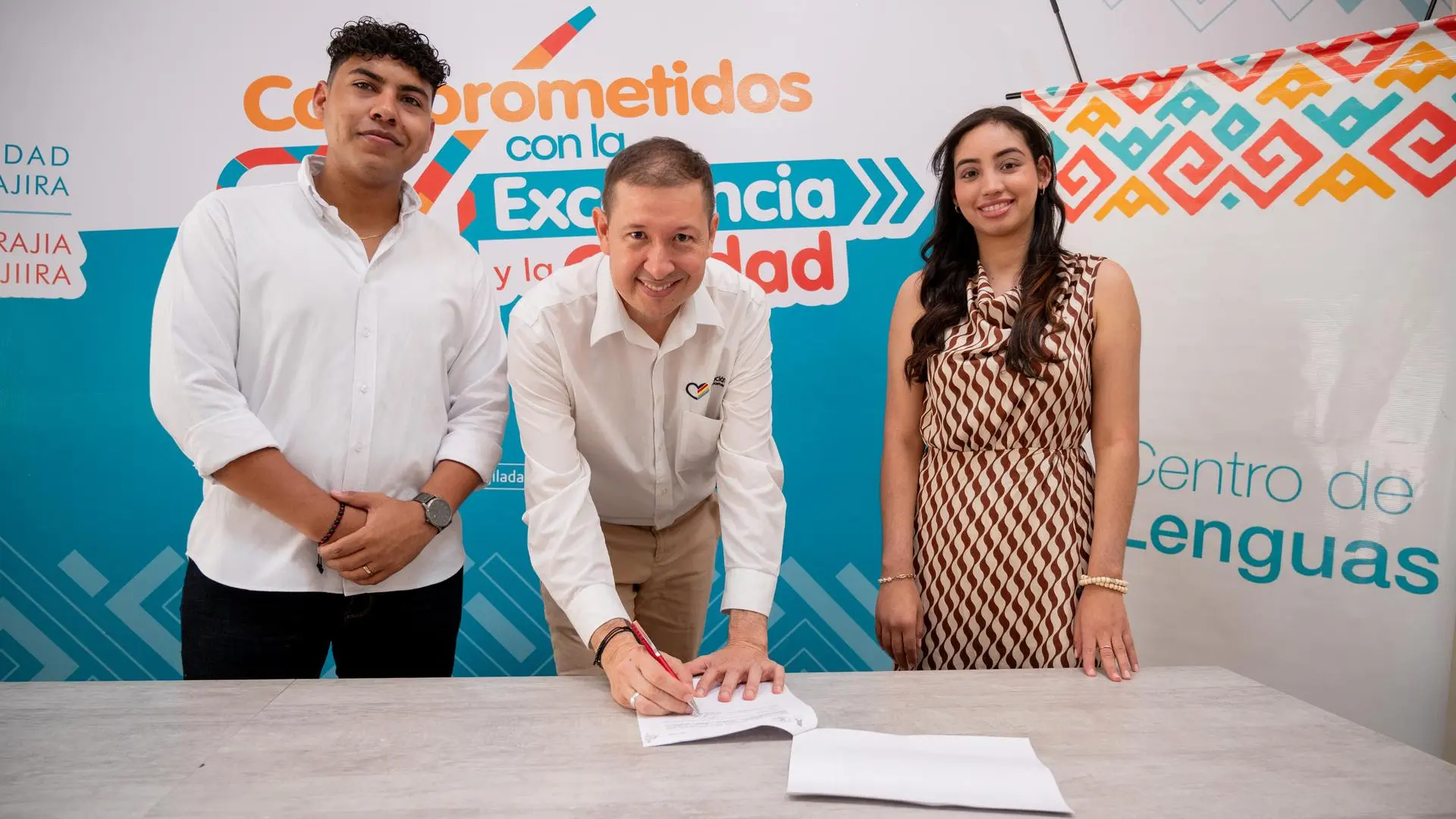 Tres personas posan sonrientes frente a un fondo institucional del Centro de Lenguas de la Universidad de La Guajira. En el centro, un hombre firma un documento sobre una mesa, mientras a su lado se encuentran un hombre y una mujer posando para la foto.
