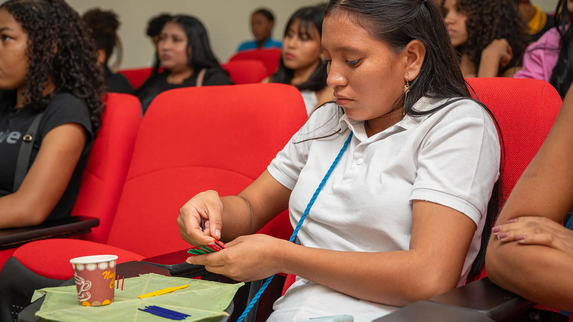 oven sentada en una silla roja durante una actividad académica o taller, concentrada en manipular palillos de colores sobre una mesa plegable. A su alrededor, otras personas observan atentamente. En el ambiente se percibe atención y participación en una dinámica práctica y educativa.