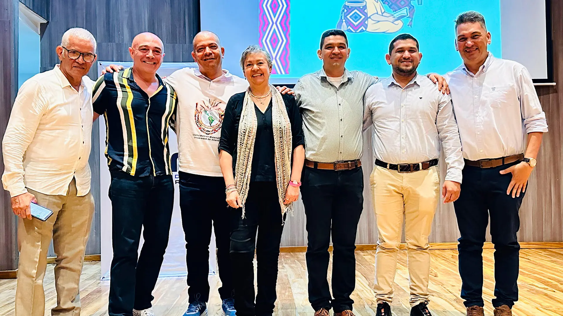 Siete personas posan sonrientes sobre un escenario de madera, frente a una pantalla con una ilustración colorida de fondo. Todos visten de manera semiformal y se encuentran con los brazos sobre los hombros, mostrando camaradería y unidad. La escena ocurre en un espacio interior con buena iluminación y ambiente académico o institucional.