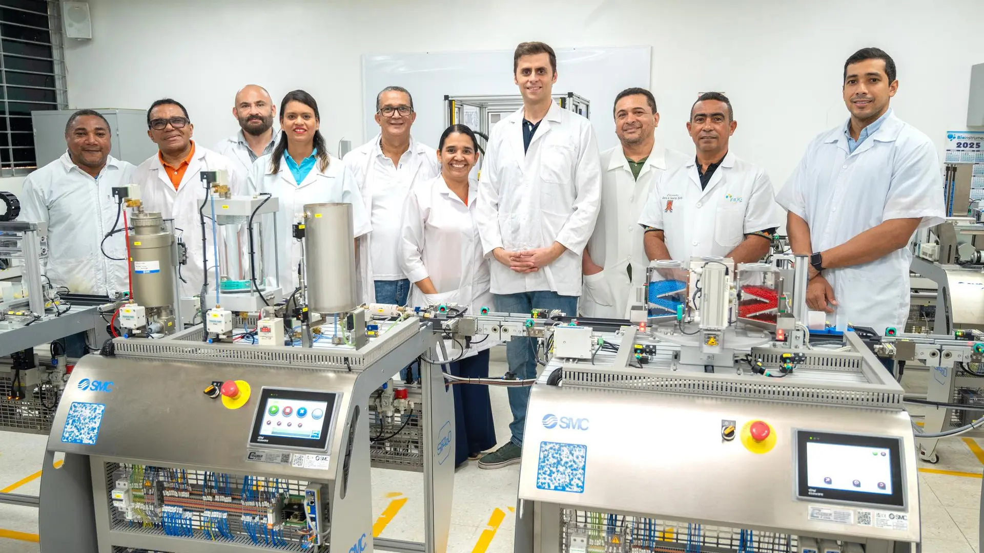 Grupo de docentes y técnicos de la Universidad de La Guajira, vestidos con batas de laboratorio, posan frente a equipos automatizados SMC en el laboratorio de automatización industrial, durante una jornada académica o de capacitación tecnológica.