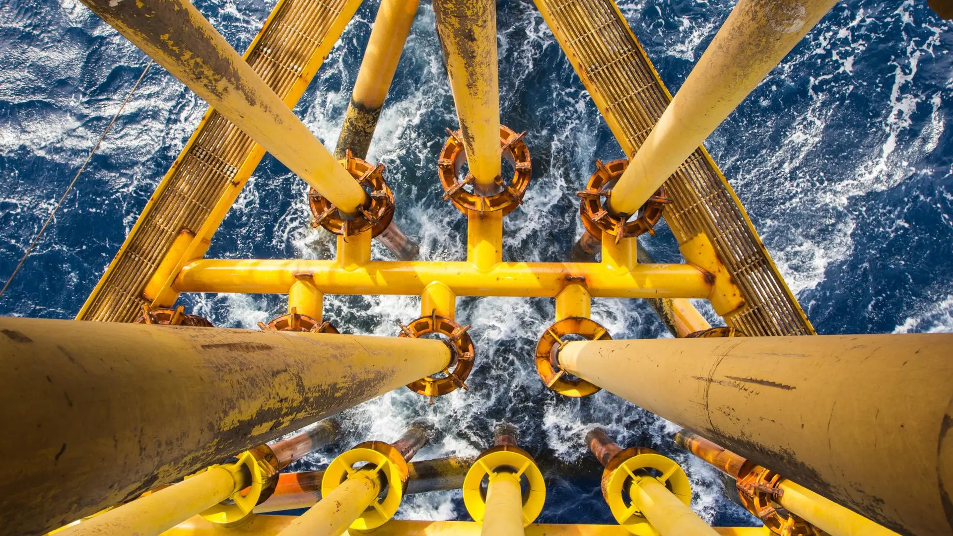 Estructura metálica amarilla de una plataforma petrolera costa afuera vista desde arriba, con tubos de perforación que descienden hacia el mar azul agitado.