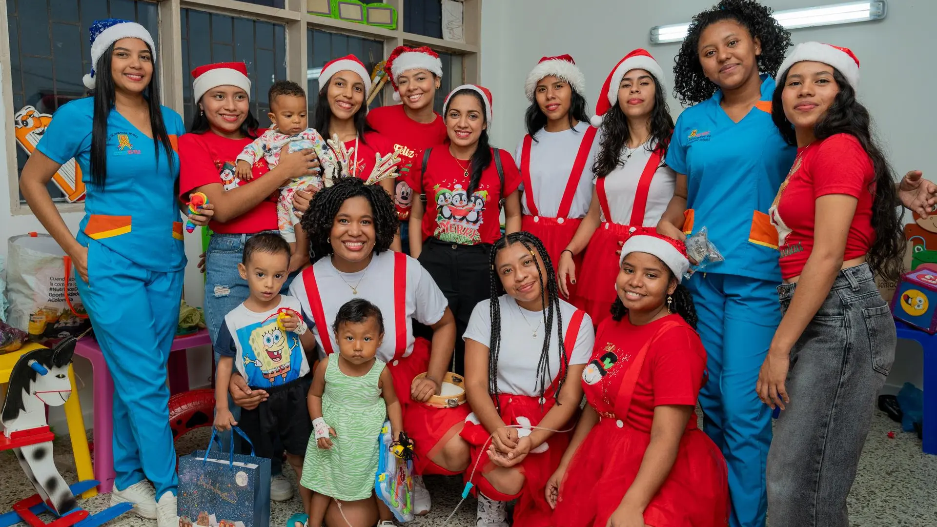 Grupo de estudiantes universitarias, algunas con gorros de Navidad y otras con uniformes azules, posan sonrientes en un salón infantil durante una actividad navideña. En el centro, dos niños pequeños acompañan a una de las estudiantes que está arrodillada. Varias jóvenes llevan camisetas rojas y faldas o tirantes navideños, sosteniendo regalos o juguetes. El ambiente es festivo y refleja el compromiso de las estudiantes con los niños y la jornada.