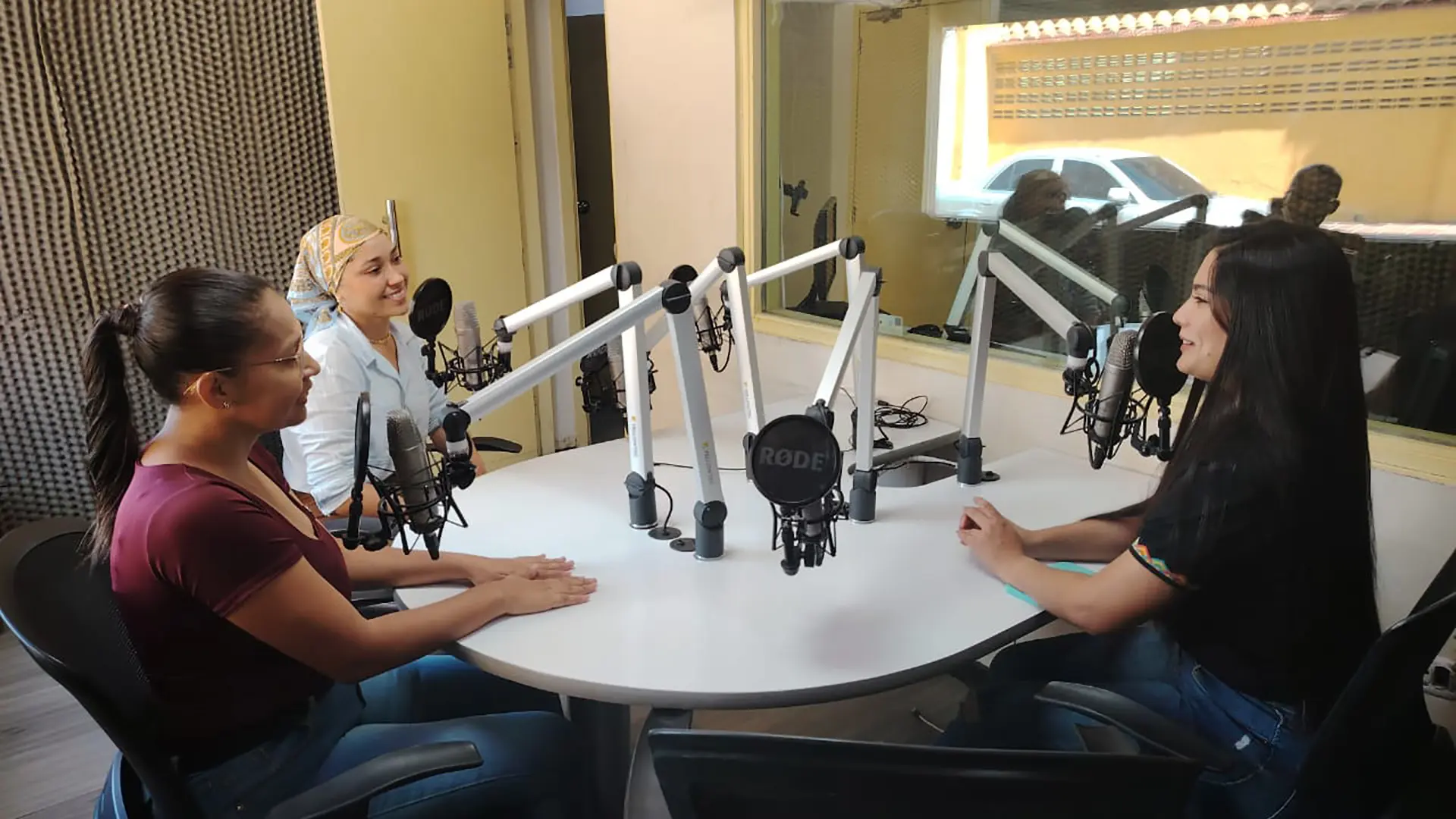 Tres mujeres participan en una conversación dentro de una cabina de radio. Están sentadas alrededor de una mesa circular equipada con micrófonos profesionales y brazos articulados. Todas sonríen mientras conversan en un ambiente ameno. A través del vidrio del estudio se puede ver el área de control y parte del exterior iluminados por la luz natural.