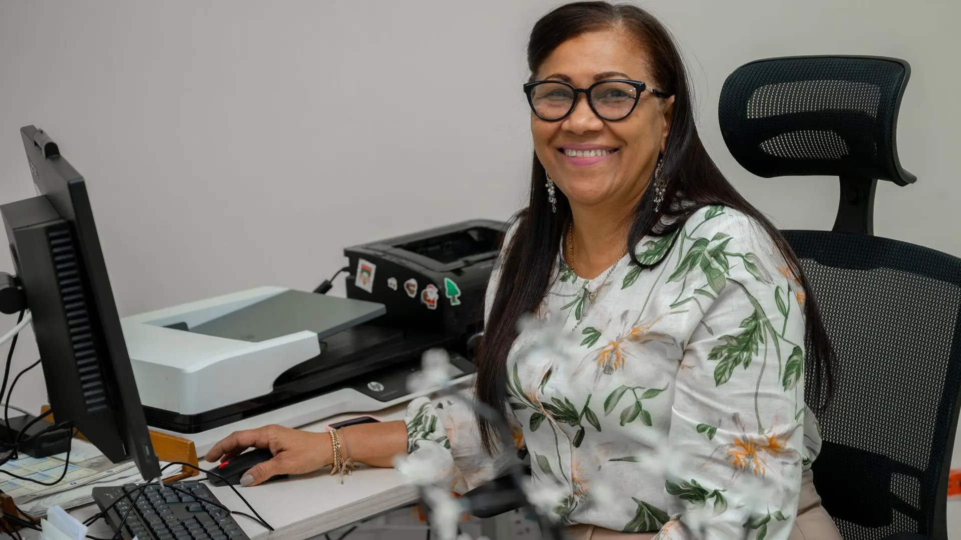 Mujer sonriente , sentada en su escritorio frente a un computador,en un entorno de oficio moderno.Viste una blusa con estampado floral y utiliza gafas,proyectando una imagen de amabilidad y profesionalismo.