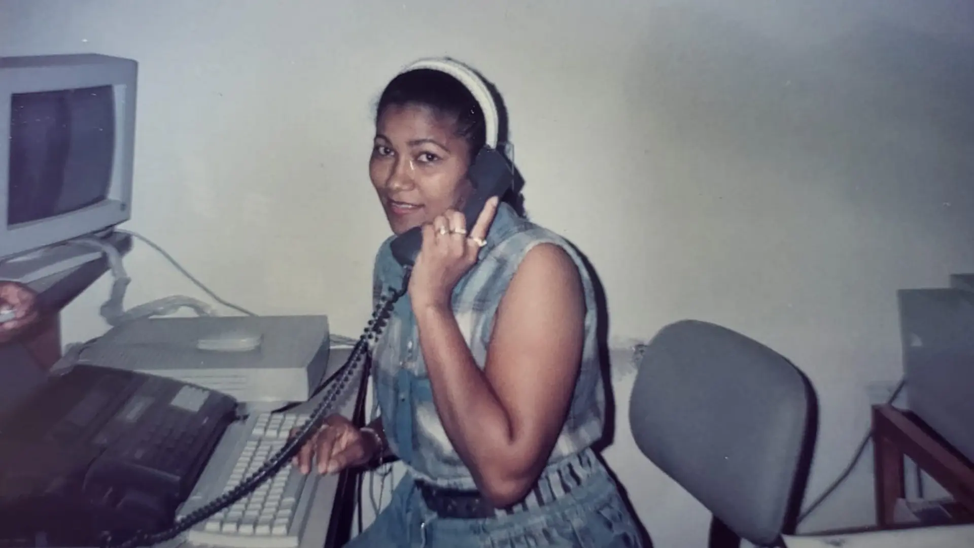 Mujer sonriente sentada en su escritorio frente a un escritorio mientras sostiene un teléfono fijo .A su lado hay un computador antiguo y una impresora,en un entorno de oficina que evoca la tecnología de los años noventa.