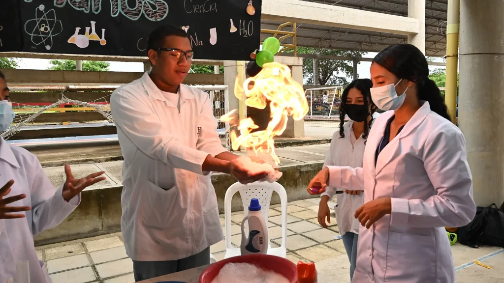 Un grupo de jóvenes con batas de laboratorio realiza un experimento químico en un espacio abierto. Un estudiante, usando gafas de seguridad, sostiene en su mano una espuma que arde en una llama controlada, mientras otra estudiante enciende el fuego con un encendedor. Otros dos observan atentos. Detrás de ellos hay un cartel decorado con dibujos de elementos científicos.