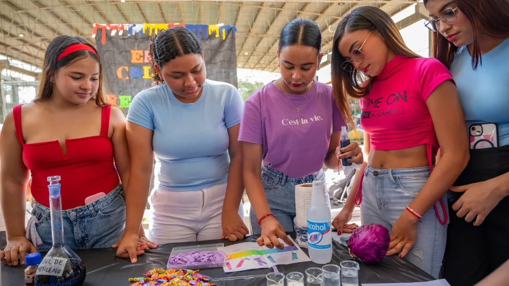 Grupo de cinco jóvenes realizando una demostración científica en una mesa cubierta con un mantel negro. Observan una serie de materiales como indicadores de pH hechos con col lombarda, pequeñas muestras líquidas en vasitos transparentes, una botella de alcohol, dulces de colores y una hoja con una escala de pH. Una de las jóvenes sostiene un frasco cuentagotas mientras otra señala la hoja de colores. Están en un espacio amplio y techado decorado con un cartel al fondo.